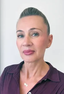 Head-and-shoulders portrait of a woman with slicked-back gray hair, wearing a burgundy blouse and a gold necklace, looking at the camera.